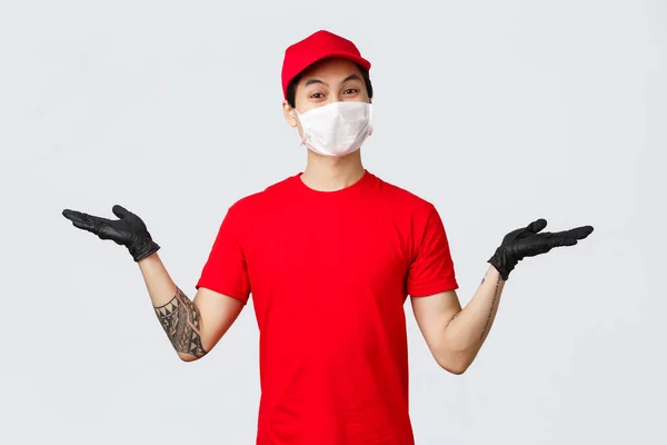 Las mejores variantes. Amistoso chico asiático repartidor sonriente en uniforme rojo, gorra y camiseta, levantar las manos para sostener los artículos, introducir productos o mensajero de seguimiento de paquetes. Concepto de compras en línea y logística —  Fotos de Stock