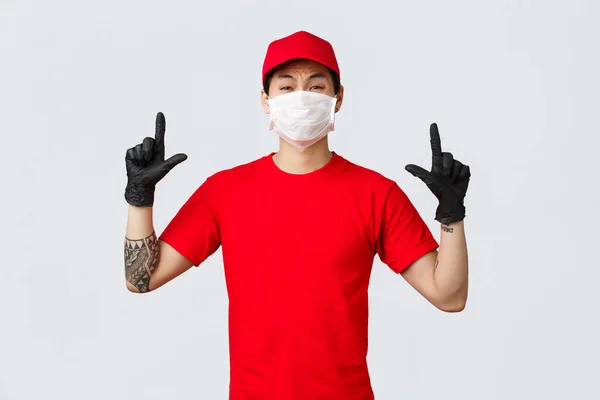 Réticent ou effrayé, gars asiatique peu sûr pleurnicher, grimaçant comme pointant les doigts vers le haut, porter uniforme de livraison, casquette rouge et t-shirt, entreprise protéger la santé du personnel avec des masques de protection et des gants — Photo