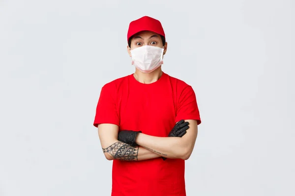 Sorprendido asiático repartidor en máscara médica y guantes de protección, vistiendo uniforme rojo, levantando las cejas asombrado con gran orden para entregar. Parece divertido. Mensajero brazos cruzados pecho —  Fotos de Stock