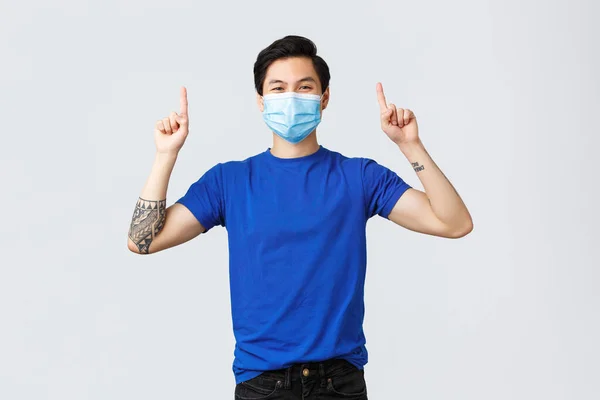 Olika känslor, social distans, självkarantän på coronavirus och livsstil koncept. Glada leende asiatisk man i medicinsk mask och t-shirt, peka fingrar upp för att annonsera, visar banner — Stockfoto