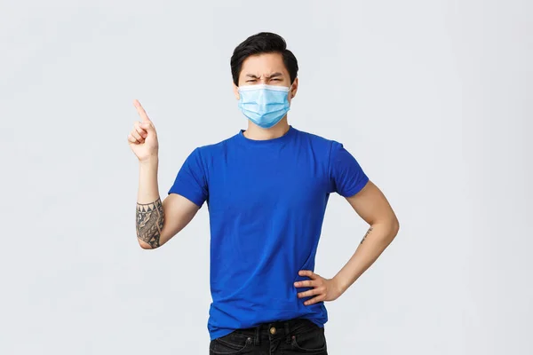 Olika känslor, social distans, självkarantän på coronavirus och livsstil koncept. Äcklig och missnöjd asiatisk man i medicinsk mask, rynkar pannan, pekar finger övre vänstra hörnet dålig sak — Stockfoto