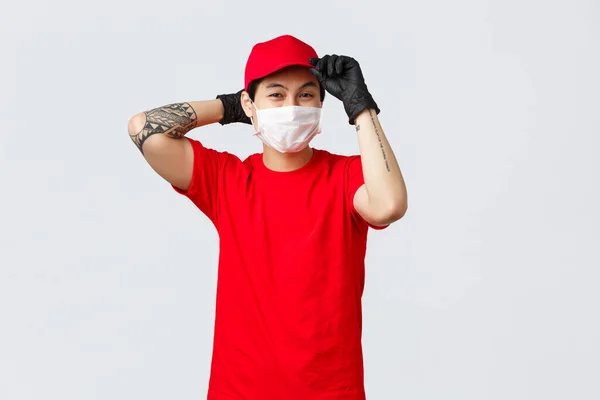 Happy young asian delivery guy put on red cap, wear medical mask and gloves, get ready deliver parcel to client, covid-19 pandemic safety measures for carriers. Courier ensure fast shipping