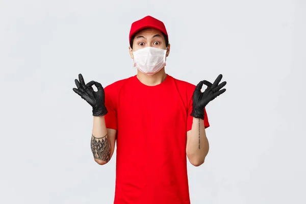 Worried young asian delivery man trying calm down nervous client, showing zen gesture, wearing medical mask and protective gloves, company uniform. Courier make okay sign ensure best quality — Stock Photo, Image