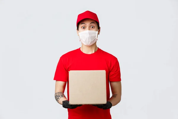 Corriere sorpreso con cappuccio uniforme rosso e t-shirt, scatola con ordine del cliente, maschera medica e guanti protettivi per la sicurezza durante il coronavirus pandemico. Uomo di consegna portare il vostro ordine — Foto Stock