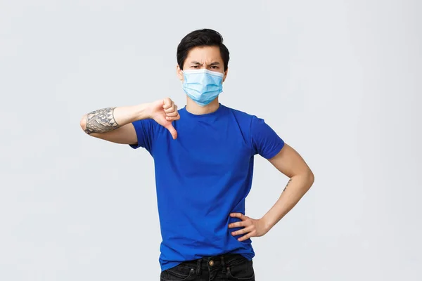 Olika känslor, social distans, självkarantän på covid-19 och livsstil koncept. Besviken och upprörd allvarlig asiatisk hane i blå t-shirt, medicinsk mask visa tummen ner — Stockfoto