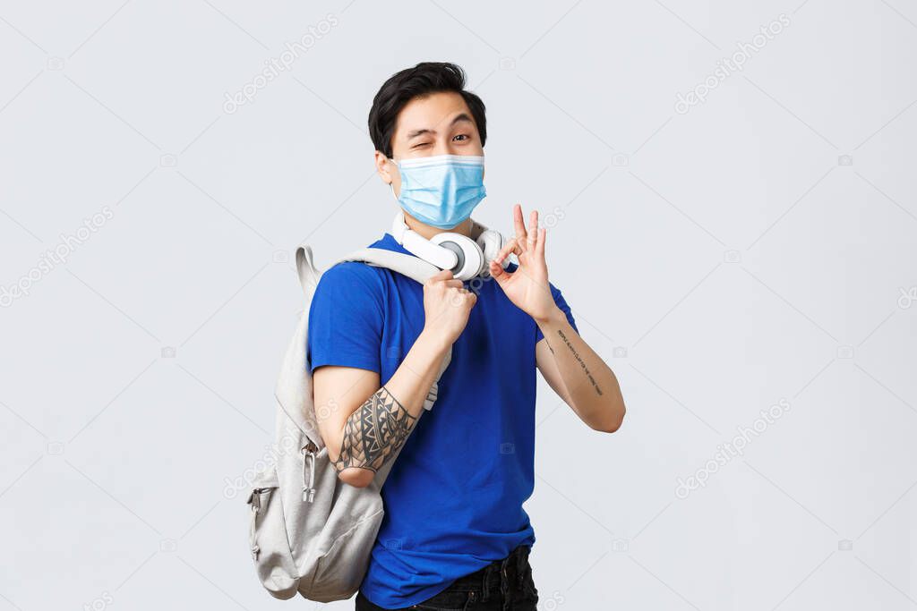 Back to school, studying on covid-19, education and university life concept. Happy handsome asian male student show okay sign, likes new year college, wink in medical mask, go classes with backpack