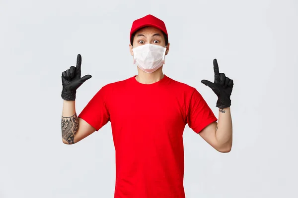 Překvapený mladý asijský doručovatel ve firemní uniformě, červené tričko a čepice, zírající kamera ohromena a šokována, ukazující prsty nahoru, nosit ochranné rukavice a lékařskou masku při práci — Stock fotografie