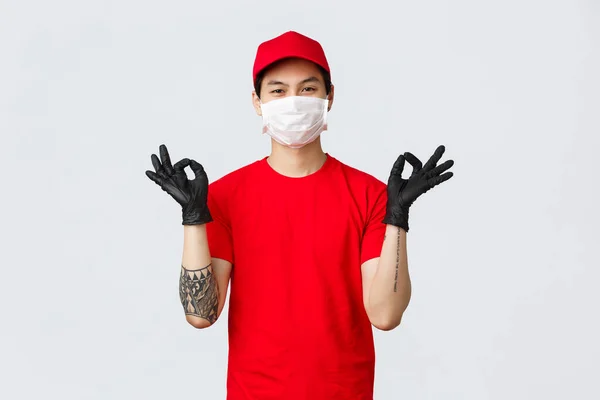 Restez calme et livrez vite. Homme asiatique souriant détendu en bonnet rouge et t-shirt, portant un masque de protection et des gants médicaux, faire preuve de geste zen, méditer. Shopping à domicile, livrer à votre porte — Photo