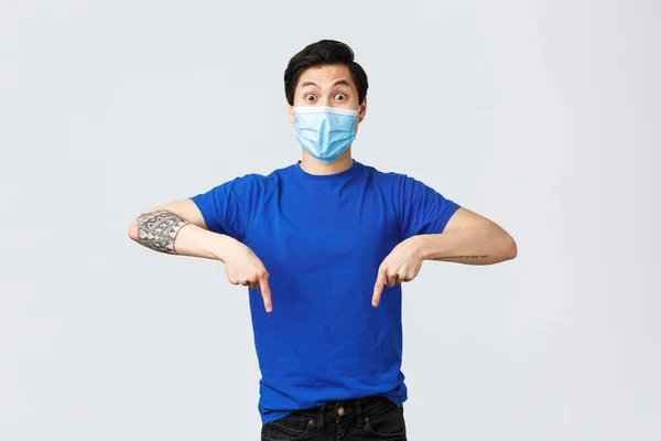 Olika känslor, social distans, självkarantän på coronavirus och livsstil koncept. Entusiastiska och upphetsad asiatisk man i medicinsk mask, pekar finger ner och ser kameran fascinerad — Stockfoto