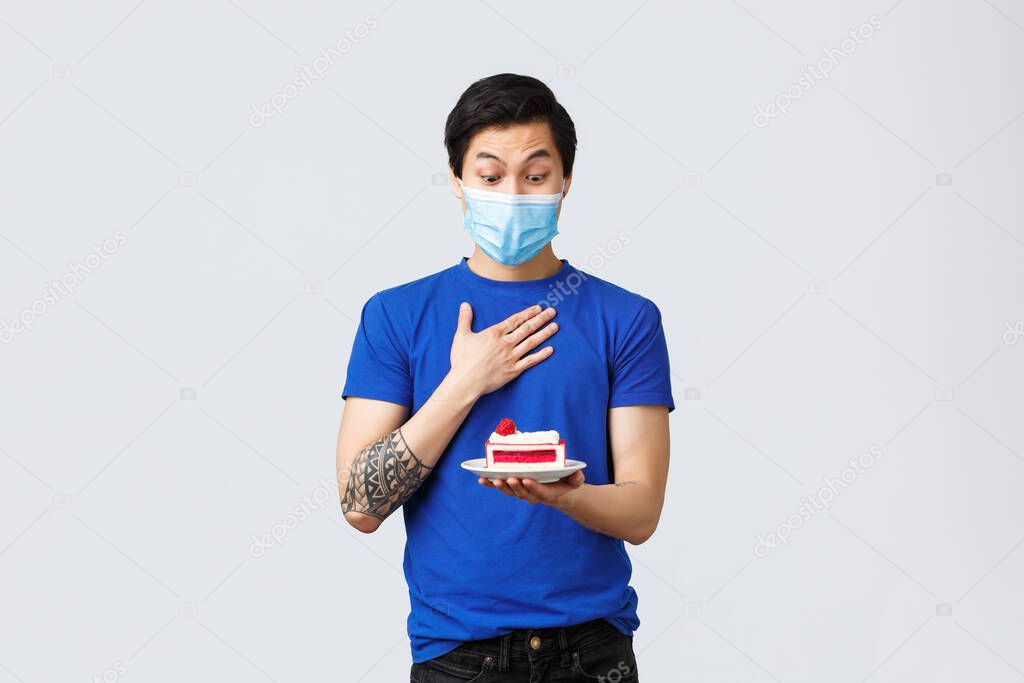 Different emotions, social distancing, quarantine on covid-19 and lifestyle concept. Happy and surprised asian guy receive cake as birthday gift, wear medical mask, look excited at dessert on plate