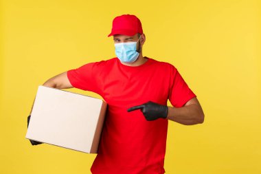 Salgın sırasında hızlı teslimat, covid-19, güvenli nakliye, alışveriş konsepti. Yakışıklı kurye paketini getir, göz kırp ve işaret kutunu, koronavirüsten koruyan tıbbi maske tak.