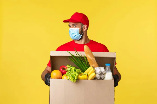 Takeaway delivery, covid-19 quarantine and groceries concept. Enthusiastic courier in red uniform, medical mask, look cheeky, bring customer food deliver, online order to doorstep