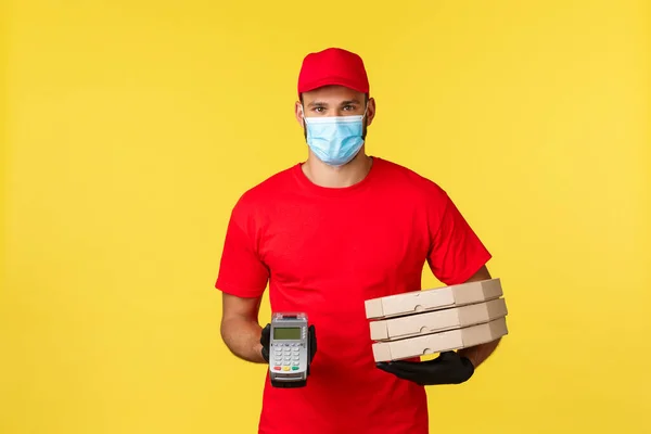 Food delivery, tracking orders, covid-19 and self-quarantine concept. Courier in red uniform cap and t-shirt, medical mask, giving client POS terminal and pizza order