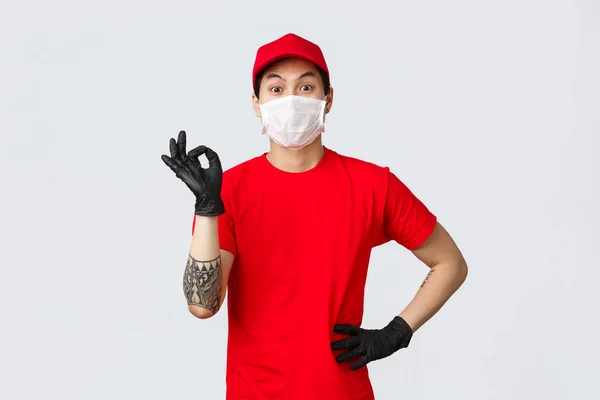 Tout va bien. Charismatique asiatique livreur gars en t-shirt rouge et casquette assurer paquet client est sûr, employé porter des masques médicaux, gants de protection lors de la pandémie covid-19, montrer ok confirmer signe — Photo