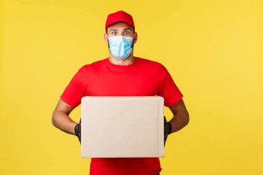 Salgın sırasında hızlı teslimat, covid-19, güvenli nakliye, online alışveriş konsepti. Kırmızı üniformalı ve tıbbi maskeli hevesli genç kurye, şaşırmış görün, müşteri sipariş kutusunu tutuyor.