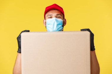 Salgın sırasında hızlı teslimat, covid-19, güvenli nakliye, online alışveriş konsepti. Tıbbi maskeli ve kırmızı şapkalı gülümseyen genç kurye, kutu kaldırıyor, sipariş paketinin yanında duruyor ve gülümsüyor.
