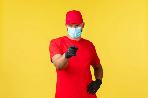 Express delivery during pandemic, covid-19, safe shipping, online shopping concept. Confident serious courier in medical mask and gloves, pointing finger at camera, yellow background — Stock Photo, Image