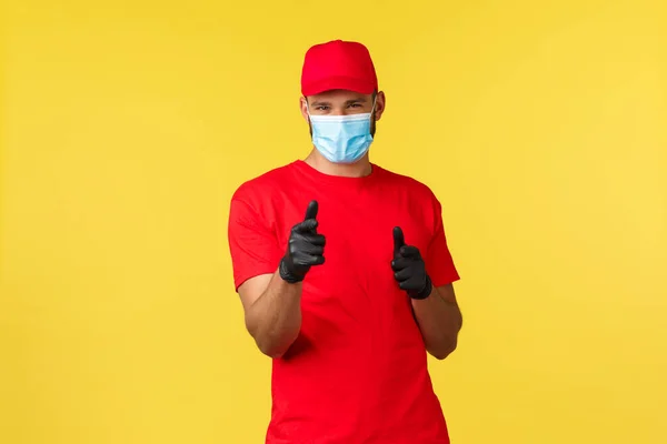 Delivery during pandemic, covid-19, safe shipping, online shopping concept. Friendly pleasant courier in red uniform, medical mask and gloves pointing cheeky camera, promote express mail — Stock Photo, Image