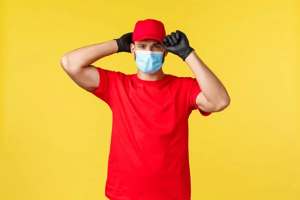 Expressleverans under pandemi, covid-19, säker frakt, online shopping koncept. Seriös stilig kurir sätta på uniform röd mössa, bära medicinsk mask och handskar, att vara redo att leverera din beställning — Stockfoto