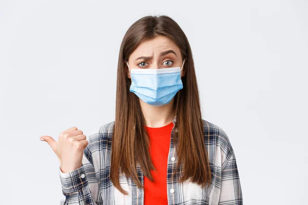 Coronavirus utbrott, fritid på karantän, social distans och känslor koncept. Skeptisk ung kvinna i medicinsk mask uttrycker misstro mot fanan till vänster, pekar tveksamt — Stockfoto