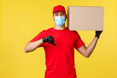 Salgın sırasında hızlı teslimat, covid-19, güvenli nakliye, online alışveriş konsepti. Kırmızı üniformalı üzgün ve hayal kırıklığına uğramış kurye, tıbbi maske, hoşnutsuzluğunu göster, paketi tut