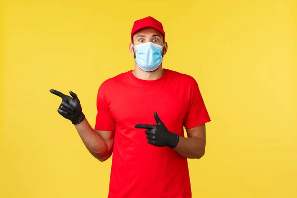 Express levering tijdens pandemie, covid-19, veilige verzending, online shopping concept. Opgewonden en onder de indruk koerier in rood uniform, medicla masker en handschoenen, wijzend naar links, hoor interessant nieuws — Stockfoto