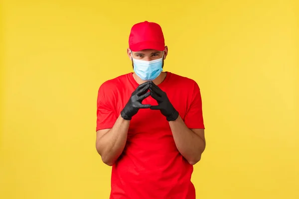Express delivery during pandemic, covid-19, safe shipping, online shopping concept. Devious and creative young courier in red uniform and medical mask, have plan, scheming over yellow background — Stock Photo, Image
