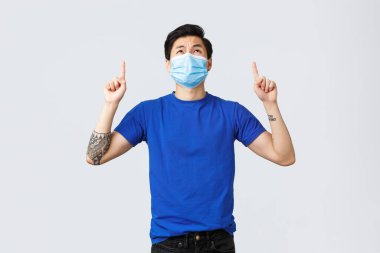 Different emotions, social distancing, self-quarantine and lifestyle concept. Disappointed and upset asian guy in medical mask and blue t-shirt, looking, pointing up with displeased grimace clipart