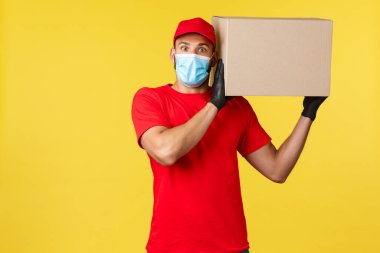 Salgın sırasında hızlı teslimat, covid-19, güvenli nakliye, online alışveriş konsepti. Şaşkın kurye kırmızı üniformalı, tıbbi maskeli, heyecanlı bir kamera gibi görün, kutuyu omzunda tut.