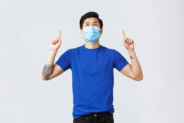 Olika känslor, social distans, självkarantän och livsstil koncept. Besviken och upprörd asiatisk kille i medicinsk mask och blå t-shirt, tittar, pekar upp med missnöjd grimace — Stockfoto