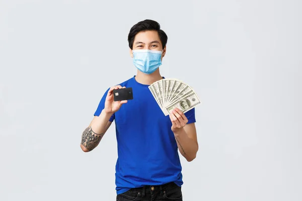 Pénz, életmód, biztosítás és befektetési koncepció. Magabiztos mosolygós ázsiai férfi javasoljuk, hogy banki és érintésmentes fizetés, bemutató hitelkártya és készpénz dollárt, viseljen orvosi maszk — Stock Fotó