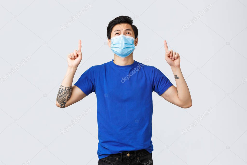 Different emotions, social distancing, self-quarantine and lifestyle concept. Disappointed and upset asian guy in medical mask and blue t-shirt, looking, pointing up with displeased grimace