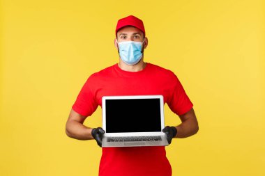 Covid-19, güvenli alışveriş, hızlı teslimat ve sipariş izleme konsepti. Kırmızı üniformalı ve tıbbi maskeli kuryenin dizüstü bilgisayarında harika bir reklam göstermesine şaşırdım ve etkilendim.
