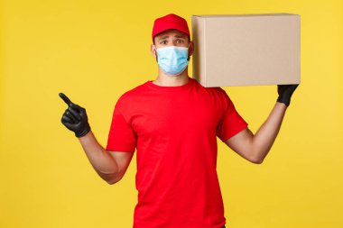 Salgın sırasında hızlı teslimat, covid-19, güvenli nakliye, online alışveriş konsepti. Üzgün ve üzgün kurye kırmızı üniformalı ve tıbbi maskeli, sol eli, kutuyu omzunda tut