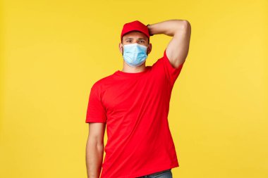Salgın sırasında hızlı teslimat, covid-19, güvenli nakliye, online alışveriş konsepti. Kırmızı üniformalı mütevazı yakışıklı kurye, tıbbi maske, kaşınan kafa, ayakta duran sarı arka plan.