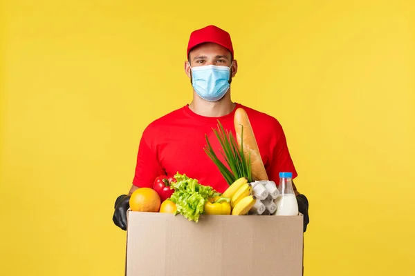 Afhaalmaaltijden, eten en drinken, covid-19 quarantaine en boodschappen concept. Vriendelijke koerier in rood uniform, medisch masker, handtas met winkelbestelling, groenten, gele achtergrond — Stockfoto