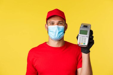 Covid-19, teslimat emirleri, alışveriş, temassız ödeme ve sosyal uzaklık konsepti. Gülümseyen yakışıklı kurye kırmızı üniformalı, tıbbi maske ve eldivenli, müşteriye POS terminali veriyor.