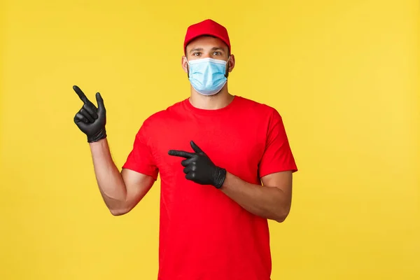 Express delivery during pandemic, covid-19, safe shipping, online shopping concept. Enthusiastic courier in red uniform, medical mask and gloves, pointing fingers left, inform about promo — Stock Photo, Image
