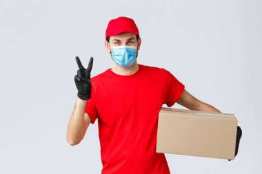 Paketler ve paket teslimatı, karantina teslimatı, nakil emirleri. Kırmızı üniformalı arkadaş canlısı kurye, yüz maskesi ve eldiven, müşteriye siparişi teslim etmek, kutuyu tutmak, barış işareti göstermek.