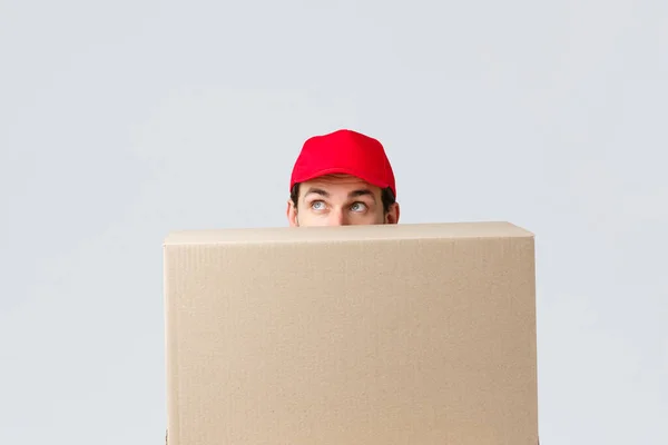 Packages and parcels delivery, covid-19 quarantine and transfer orders. Cute courier in red uniform cap hiding behind large box order, looking up intrigued, peeking over grey background — Stock Photo, Image