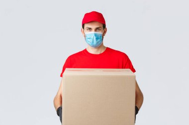 Erzak ve paket teslimatı, covid-19, karantina ve alışveriş konsepti. Kırmızı üniformalı ciddi bir kurye, eldivenler ve koruyucu yüz maskesi, Coronavirus sırasında müşteri evine paket kutusu teslim etmek.