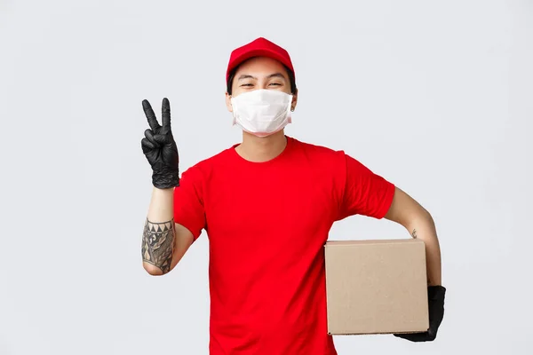Έννοια του τοκετού, πανδημία του κορωνοϊού. Χαρούμενη χαμογελαστή ασιατική courier σε κόκκινο καπέλο, t-shirt, δείχνουν την ειρήνη σημάδι, κρατώντας δέμα, φορούν ιατρική μάσκα και γάντια για την ασφάλεια των πελατών, παραδώστε το πακέτο — Φωτογραφία Αρχείου