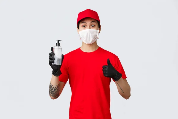 Szórakoztató futár piros sapkában és pólóban, hüvelykujj felirat, védőkesztyű és orvosi maszk viselése, az ügyfelek és a cég dolgozói kézfertőtlenítőt kapnak. Az érintésmentes biztonságos szállítás koncepciója — Stock Fotó