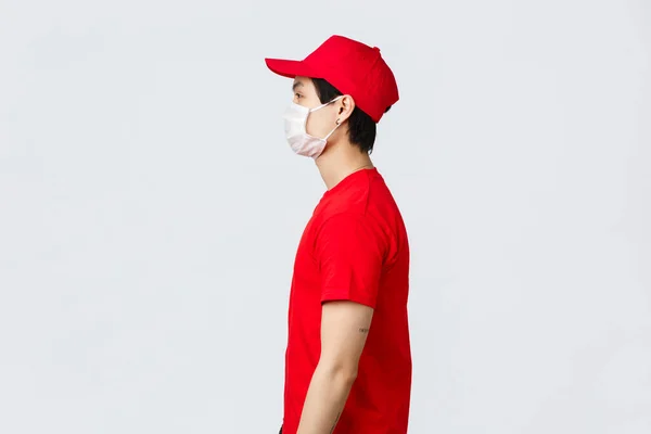 Profilbild eines asiatischen Zustellers in roter Mütze und T-Shirt, mit medizinischer Schutzmaske, Blick nach links, Warten vor der Haustür auf das Abholpaket des Kunden, Kurierdienst während der Pandemiezeit 19 — Stockfoto