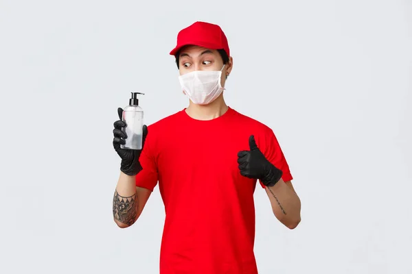 Curioso asiático homem de entrega uniforme vermelho, máscara médica e luvas de proteção recebeu higienizador de mão da empresa transportadora, mostrar thumb-up, impressionado como o empregador cuidar de seus funcionários durante covid 19 — Fotografia de Stock