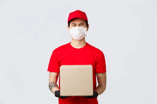 Angry asian delivery man in red cap, t-shirt, holding box, wear medical mask and gloves, complaining on rude client, frowning aversion and dislike. Courier frowning disappointed