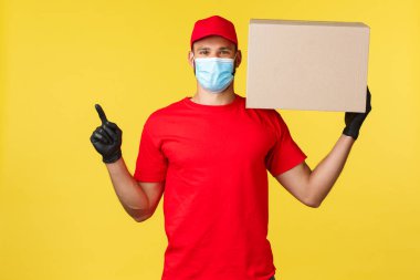 Salgın sırasında hızlı teslimat, covid-19, güvenli nakliye, online alışveriş konsepti. Yakışıklı teslimatçı, paketli kurye kutusu sol üst köşeyi işaret ediyor, tıbbi maske takıyor.