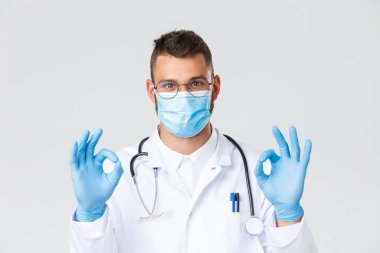 Covid-19, sağlık çalışanları, salgın hastalıklar ve virüs önleme konsepti. Beyaz önlüklü ve gözlüklü yakışıklı doktor, sağlık maskeli doktor tamam işareti, tanıtım önerisi.