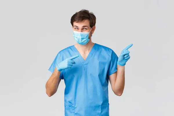 Covid-19, gezondheidswerkers en ziekenhuisconcept. Enthousiaste arts in medisch masker, handschoenen en blauwe verpleegkundige of chirurg schrobt wijzende vingers naar rechts, wijzend weg, reclame promoten — Stockfoto