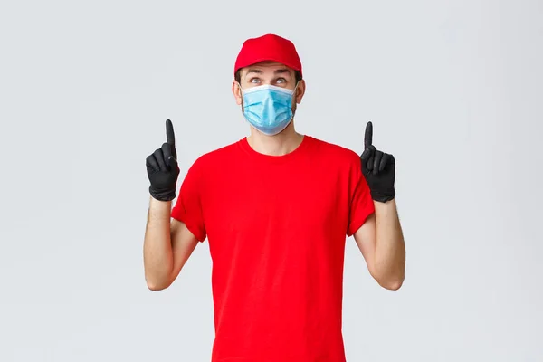 Covid-19, zelf-quarantaine, online winkelen en verzending concept. Lachende bezorger rood uniform pet en t-shirt, medisch masker met rubberen handschoenen, op zoek naar nieuwsgierig, leesbord of promo — Stockfoto